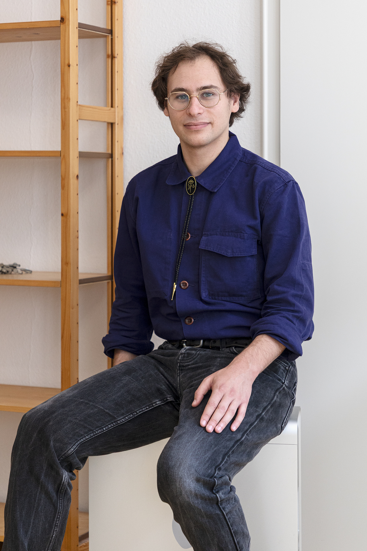 Bernd Seeland Junger Mann sitzt auf einer weißen Möbelkante, trägt blaue Hemdbluse und dunkle Jeans.