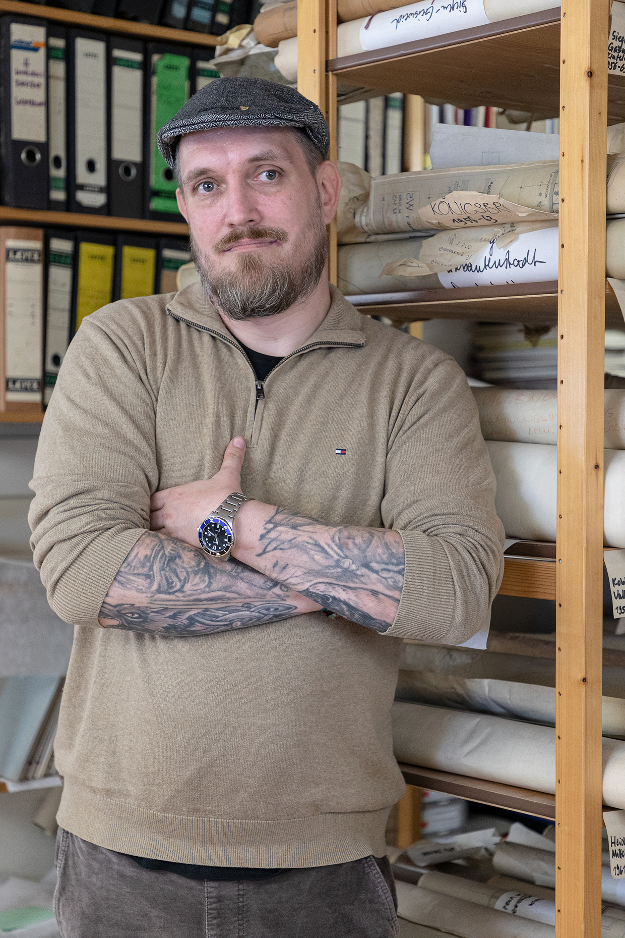 Bernd Seeland Mann mit Tattoos und Uhrenarmband, beige Pullover, Arme verschränkt, Regal mit Rollenrollen im Hintergrund.